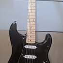 Černá elektrická kytara typu Stratocaster se světlým hnedem hmatníkem a bílým pickguardem. Na hlavě krku je viditelné logo Fender. Nástroj má tři bíl…