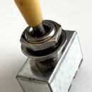 Kovový přepínač s béžovou plastovou knoflíkovou koncovkou. Tělo je vyrobeno z matného stříbrného kovu s viditelnými škrábanci a stopami oxidace v dol…