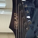 Černá hlava elektrické kytary s nápisem SOLAR GUITARS. Na hlavě jsou vidět šest ladicích kolíčů a struny. Povrch je v matném černém provedení bez vid…