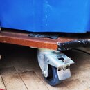 Detailní pohled na spodní část modrého objektu s dřevěnou základnou. Nástroj je osazen kovovým otočným kolečkem s gumovou bandaží. Na hraně dřevěné d…