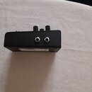 Černý efekt pedál v kovovém provedení. Na zadní straně jsou dva vstupní/výstupní jack konektory a nahoře dvě černév knoflíky. Dole je viditelný bílý…