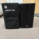 Na fotografii je černý subwoofer a přiléhající černý transportní kryt z látky. Na krytu je bílé logo „HK AUDIO“ a nápis „LINEAR SUB“. Subwoofer má če…