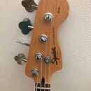 Fotografie zachycuje hlavu basové kytary z přírodního dřeva s čtyřmi kovovými ladičkami. Na povrchu je černý nápis Squier by Fender a PRECISION BASS®…