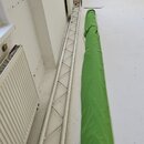 Na fotografii je stříbrný hliníkový mřížový truss v poloze vleže na podlaze. Vedle něj leží dlouhý zelený textilní kryt nebo přepravní vak. Na vybave…