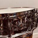 Detailní pohled na chromovaný snare drum s bílým blátcem. Nástroj má lesklý kovový povrch a viditelné chromované lugy. Na korpusu nejsou žádná čiteln…