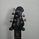 Fotografie zachycuje hlavu černé elektrické kytary s šesti stříbrnými ladičkami. Na povrchu je bílý nápis „Sterling by MUSIC MAN“ a stylizovaný podpi…