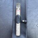 Kondenzátorový mikrofon stříbrné barvy s mřížkou v horní části a červeným prstenem pod ní. Mikrofon je umístěn v černém plastovém držáku s logem znač…
