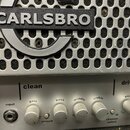 Detailní pohled na přední panel kytarového komba. Na stříbrné kovové mřížce je umístěno černé logo s bílým nápisem CARLSBRO. Spodní ovládací panel ob…
