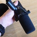 Černý dynamický mikrofon s nasazenou větrolamem z pěny, upevněný v držáku na stojanu. Na těle mikrofonu je bílý nápis SHURE SM7B. Vedle mikrofonu lež…