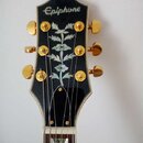 Fotografie zachycuje hlavu kytary v černé barvě se zlatými ladičkami. V horní části je bílý nápis Epiphone a uprostřed se nachází velká perleťová int…