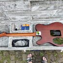 V otevřeném hnědém tvrdém kufru leží elektrická kytara s hnědým dřevěným povrchem a černou plastovou krytkou elektroniky. Na krku kytary je vidět kov…