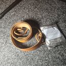 Hnědý kožený řemen s copánými konci. Vedle něj je stříbrné balení strun s nápisy CORROSION PROTECTOR a D'Addario. V horní části balení jsou viditelné…