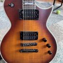 Elektrická kytara s trupem v hnědém sunburst provedení a viditnou strukturou dřeva. Nástroj má dva černá humbuckery, tři černé oválné ovládací knofík…