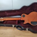 Oranžová elektrická kytara v černém tvrdém kufru s červeným plyšovým vnitřkem. Na hlavě nástroje je bílý nápis Gibson. Povrch kytary je glossy, s čer…