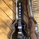 Černá elektrická kytara typu Les Paul v tvrdém kufru s šedým polstrováním. Na hlavě hmatníku je bílý nápis Epiphone. Kytara má dva humbuckery, stříbr…