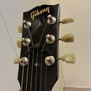 Fotografie zachycuje hlavu černé elektrické kytary se zlatým logem Gibson. Nástroj má šest ladicích kol s krémovými plastovými knoflíky. Na povrchu j…