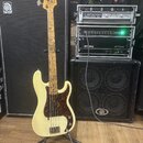 Krémově bílá elektrická basová kytara s hnědým vzorovaným pickguardem a javorovým hnedým hmatníkem. Na hlavě nástroje je viditelné logo Fender. Kytar…