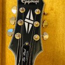 Černá hlava elektrické kytary s bílým lemováním a zlatými ladičkami. V horní části je bílé logo Epiphone, uprostřed je bílý kosočtvercový ornament a…