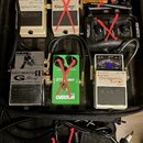 V černé brašně je několik kytarových efektů a příslušenství. Jsou zde pedály značky BOSS s nápisy Digital Delay DD-3, Equalizer GE-7 a Chromatic Tune…