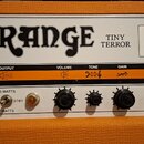 Detail kytarového zesilovače s oranžovým povrchem a bílým předním panelem. Na panelu je velký černý nápis ORANGE a vedle něj text TINY TERROR. Pod ní…