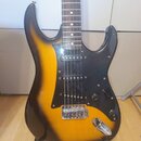 Elektrická kytara s trupem v černo-oranžovém sunburst provedení a černým pickguardem. Nástroj má tři snímače typu single-coil, jeden tónový potenciom…