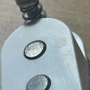 Detailní záběr na bílý kovový díl s dvěma kulatými otvory, ve kterých jsou vidět kovové součástky. Na povrchu je vpravo vyražené logo ve tvaru trojlí…
