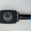 Černý dynamický mikrofon s obdélníkovou mřížkou. Na těle je bílý nápis SENNHEISER e906 a logo značky. Na zadní části těla je nalepený modrý pás s ruč…