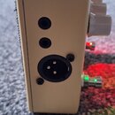 Detailní pohled na boční stranu béžového efekt pedálu. Jsou vidět dva malé vstupy jack a jeden větší konektor XLR s pěti piny. Na pravé straně jsou v…