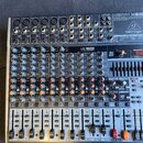 Míchací pult značky behringer, model XENYX X1822USB. Zařízení je v kovovém šasi s množstvím regulačních knoflíků, posuvníků a konektorů. Na horní čás…