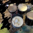Sada bubnů v tmavě červené barvě s kovovými obručemi. Na blanách jsou vidět loga EVANS a stopy po lepeních. Povrchy blan vykazují výrazné opotřebení…