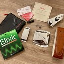 Sada příslušenství k hudebnímu nástroji rozložená na dřevěném povrchu. Obsahuje černý textilní obal s logem Fender, hnědý kožený řemen s vyraženým lo…