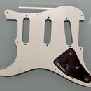Bílá pickguard pro elektrickou kytaru s třemi výřezy pro snímače. V pravé dolní části je připevněn tmavě fialový nebo černý plastový kryt s otvory pr…