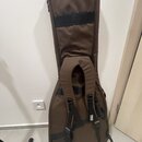 Hnědý polstrovaný obal na kytaru z textilního materiálu. V dolní části je vidět bílý nápis PRS. Obal je vybaven černými popruhy s plastovými přezkami…