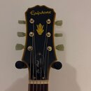 Fotografie zachycuje hlavu elektrické kytary s černým povrchem a zlatým lemováním. V horní části je nápis Epiphone a uprostřed se nachází zlatý symbo…
