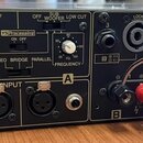 Zadní panel černého výkonového zesilovače s bílými nápisy. Jsou zde vidět ovládací prvky jako přepínače STEREO, BRIDGE a PARALLEL, dva otočné knoflík…
