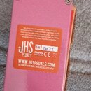 Spodní strana růžového efekt pedálu z kovu. Na spodní straně je bílý nápis WWW.JHSPEDALS.COM a text DESIGNED AND BUILT IN KANSAS CITY, USA. Je zde lo…