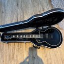 Černá elektrická kytara v tvrdém kufru s černým vnitřkem. Na hlavě hmatníku je bílé logo "Gibson". Kytara má dva humbuckery, čtyři ovládací knofíky a…