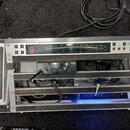 Hliníkový rack s několika zařízeními a bílým pedálem vlevo. V horní části je digitální tuner s nápisem DTR-1 DIGITAL TUNER, v prostřední části černý…