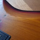 Detail zadní strany korpusu elektrické kytary s dřevěnou strukturou a barevným přechodem od žluté přes červenou k černé. V horní části jsou vidět čty…