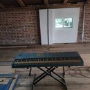 Modrý elektronický klavír umístěný na černém X-stojanu. Nástroj má standardní klávesnici a ovládací panel s tlačítky a posuvníky. Na povrchu nejsou v…