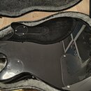 Černá elektrická kytara v otevřeném kufru s šedým plyšovým vnitřkem. Fotografie zachycuje zadní stranu trupu s viditelnými šrouby u otvorů pro zapoje…