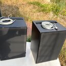 Dva černé, hranolité reproduktory umístěné na bílém stole. Na horní části každého reproduktoru leží smotaný bílý kabel. Povrchy jsou matně černé s vi…