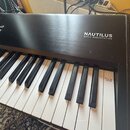 Černý digitální klavír s nápisem NAUTILUS MUSIC WORKSTATION na horním panelu. Na pravé straně klávesnice je viditelný červenobílý štítek s textem MAD…