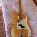 Elektrická basová kytara s přírodním dřevěným povrchem těla a hmatníkem. Nástroj má bílý pickguard, dva černé snímače a dvě ovládací knoflíky. Na hla…
