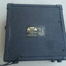 Zadní strana černého kytarového komba potaženého texturovaným vinylen. Uprostřed jsou dvě stříbrné štítky s nápisy „CAUTION“, „CE“, „INPUT POWER 220/…