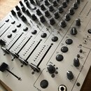 Detailní pohled na ovládací panel stříbrného mixážního pultu s množstvím černých a šedých otočných knoflíků. Na panelu jsou vidět čtyři posuvníky a r…