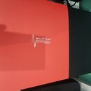 Fotografie zobrazuje dvě čtvercové plochy, jedna je červená a druhá černá. Na červené ploše je bílý nápis s logem znázorňujícím kmitající linku a tex…