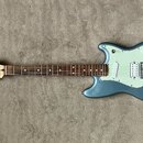 Modře šedá elektrická kytara s bílým pickguardem a hnědým hmatníkem. Na hlavě je viditelné logo "Fender". Nástroj má konfiguraci snímačů dva jednoduc…