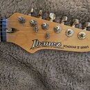 Fotografie zachycuje hlavu elektrické kytary z přírodního dřeva se šesti zlatými ladičkami. Na dřevěné části je černý nápis Ibanez a pod ním menší te…