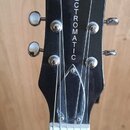 Černá hlava elektrické kytary s chromovanými ladičkami. V horní části je bílý nápis GRETSCH a svisle pod ním nápis ELECTROMATIC. Povrch je hladký, be…